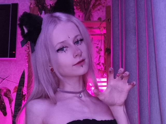 ExcitedBlizzard with pale skin, blonde hair, black cat ears, makeup, nipple piercing, choker, and painted nails, holding up a hand, pink neon lighting, smiling.