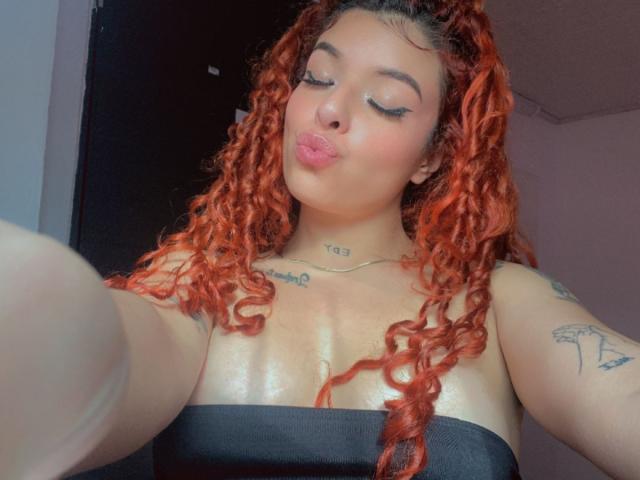 TammyyRoss with red curly hair, puffy lips, and tattoos, wearing a black strap top, taking a selfie, showing shiny skin and small tits.