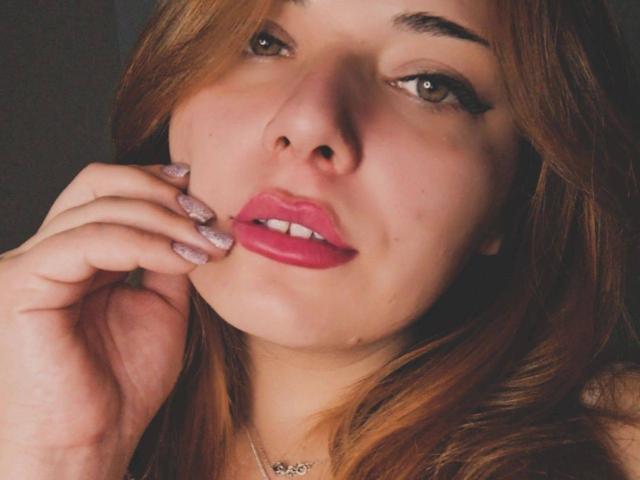 LolaFoxiX ha il rossetto rosa, le unghie scintillanti e i capelli castani ondulati, e tocca la guancia con un sorriso carino e leggermente aperto. I suoi occhi castani sono rivolti verso la telecamera..
