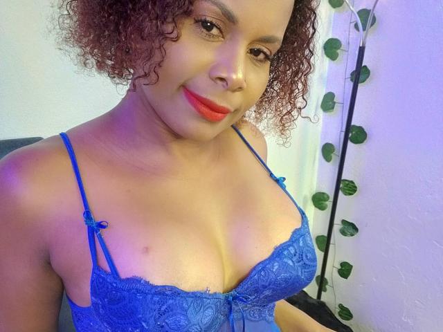 Afrodita Kane mit lockigem, braunem Haar, heller Hautfarbe, rotem Lippenstift und einem blauen Spitzen-BH, der ihre Brüste zeigt und eine kleine Nippel-Piercing, lächelnd..