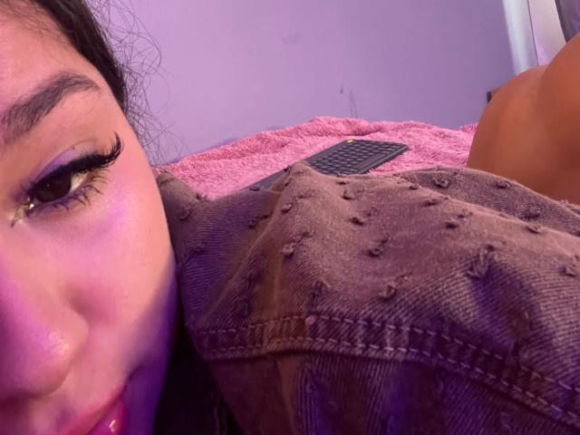 AlessiaFerry's face with thick eyelashes, purple eyeshadow, and pink lipstick, leaning on a brown, textured, button-up shirt. Her dark hair is partially visible. Her nipple is slightly exposed.