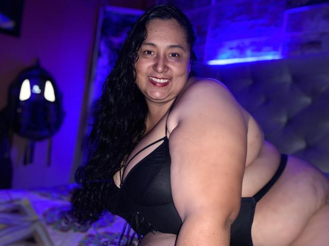TistMature with large, curvy body and black bra, smiling at camera. She has long, wavy black hair, and is leaning forward, showing her big ass and full tits.