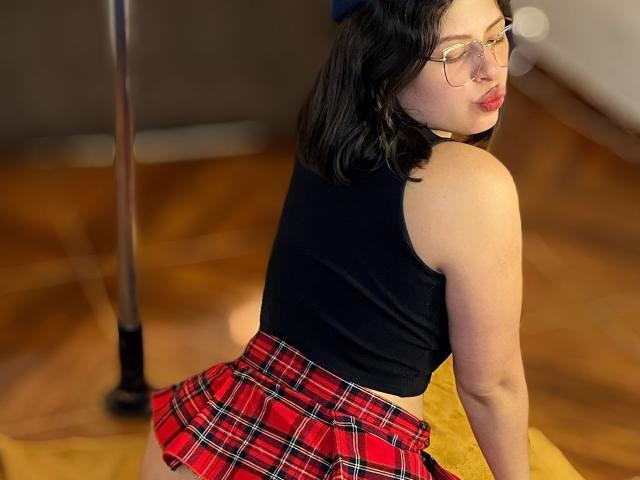 ShyVibes with glasses, black sleeveless top, red plaid skirt, and pouty lips, kneeling on wooden floor, showing her ass, black hair, and fair skin.