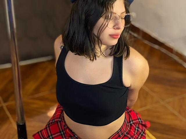 ShyVibes con cabello corto negro, gafas, top corto negro, falda escocesa roja y busto visible, sentada en el suelo de madera, mirando de lado, sosteniendo un palo frente a ella..