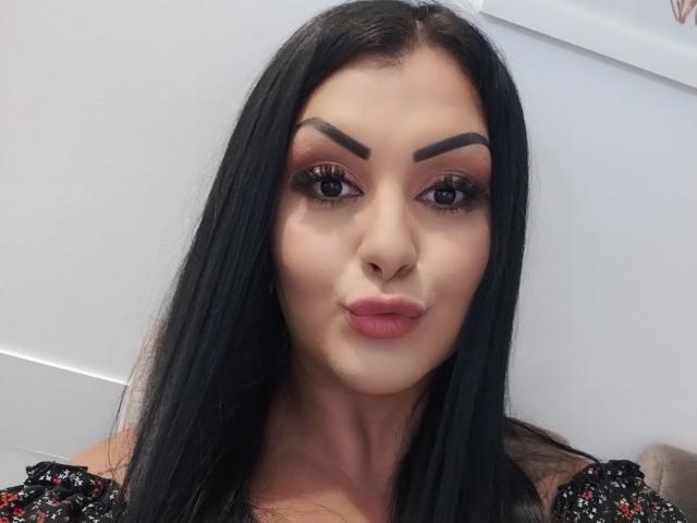 JulianaFenix has black hair, thick eyebrows, and pink lipstick with full, pouty lips. She's wearing a black, sparkly top with red accents. Her eyes are heavily lined.