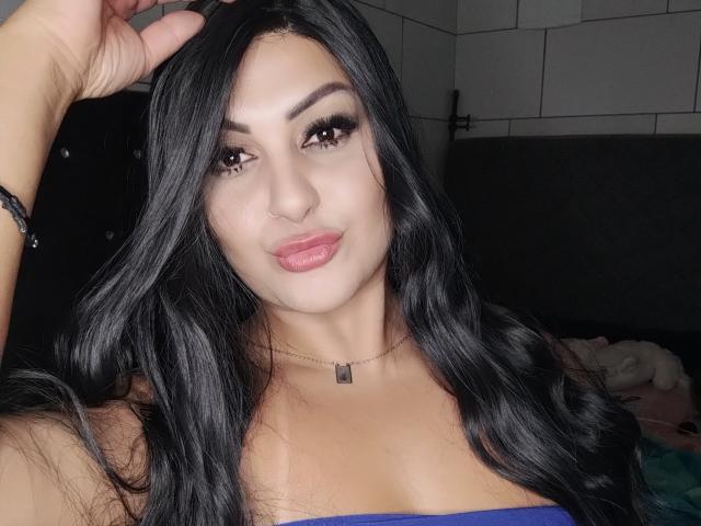 JulianaFenix has long black hair, a nose ring, and is wearing a blue top, showing her tits. She has a necklace, full lips, and a bracelet on her right hand.