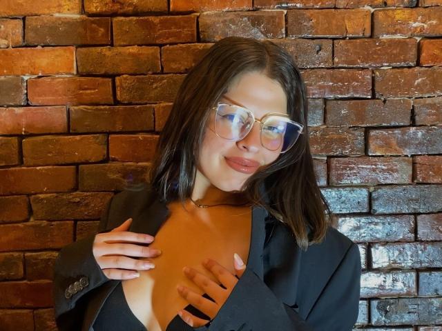 ShyVibes with glasses, black shirt, and hand on tits, smiling at camera, brown brick wall in background, wearing a gold necklace, medium-length brown hair, reflective glasses.