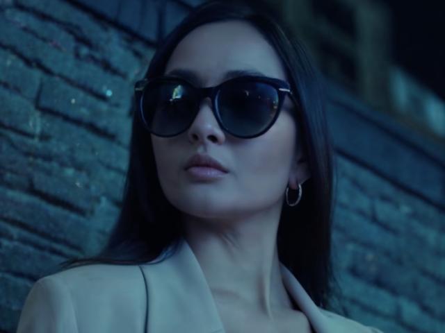 Dionisia with dark sunglasses, long brown hair, pale skin, hoop earrings, and a beige jacket, looking serious, blue-tinted, night-time, reflection in glasses.