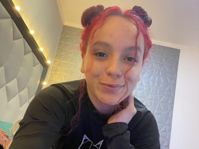 MaiaPark with red hair in double buns, black shirt, and a slight smirk, resting her chin on her hand, showing a small zit on her forehead.