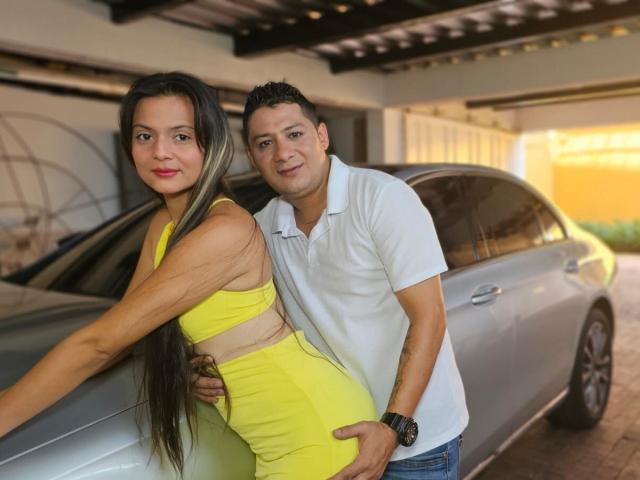 ValentinaAndTerry with long brown hair, yellow dress, and tits, leaning on a car. Terry, in a white polo, stands behind her, hand on her yellow dress-covered ass.