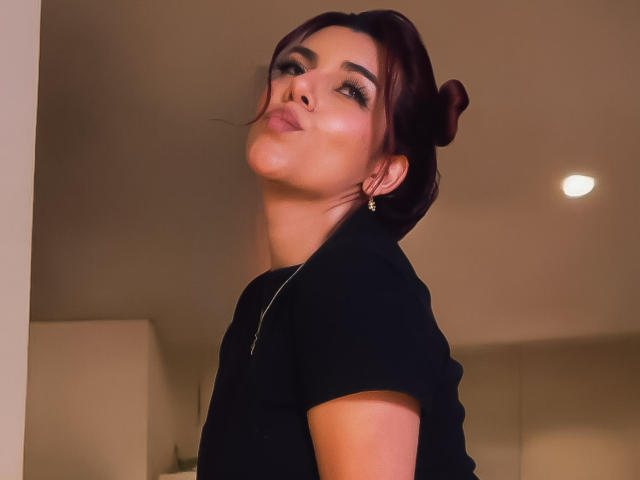 ZoeeMar, con cabello castaño oscuro recogido en un moño, camisa negra, labios fruncidos y una ligera inclinación hacia arriba, con pequeños aretes, con una expresión de suficiencia y una sonrisa sutil..