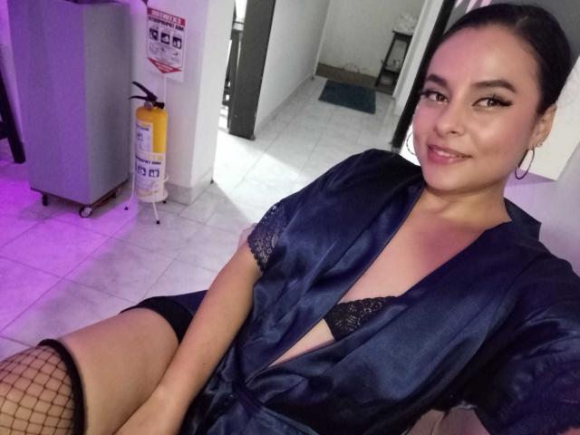 LunaSoler with dark hair, wearing a navy blue silk robe, black lace bra, and fishnet stockings, sitting, showing her tits, smiling, in a room with purple lighting.