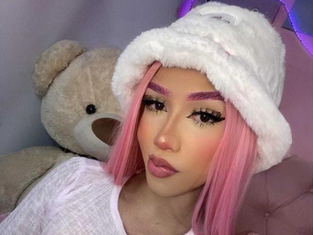 AlissLovely with pink hair, wearing a white fluffy hat, and a white shirt, has thick eyebrows, long eyelashes, and pink lipstick, standing next to a large beige teddy bear.