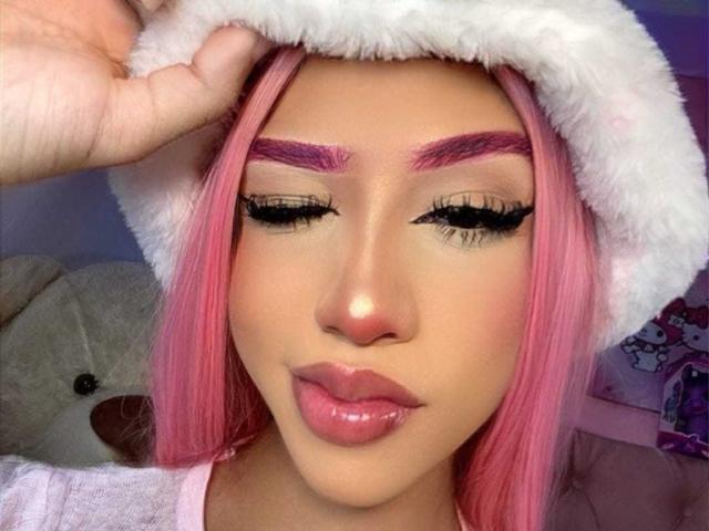AlissLovely with pink hair, white furry hat, purple eyebrows, long black eyelashes, glossy pink lips, and a hand touching her hat. Boobs not visible. Soft, pretty face.