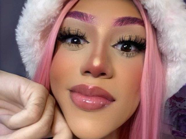 AlissLovely with pink hair, purple eyebrows, long eyelashes, glossy pink lips, wearing a white furry hat, holding her chin, looking at the camera, with a cute smile.
