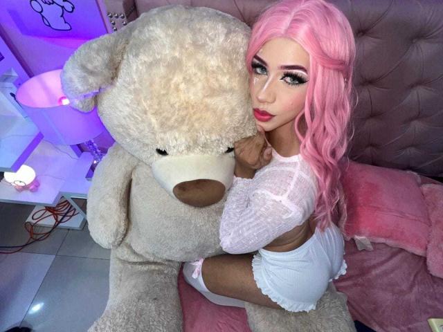 AlissLovely with pink hair, white lingerie, and thigh-high socks, snuggles against a huge beige teddy bear. Her big ass is visible, and she has long, curled eyelashes.