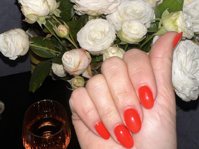 Trima's hands with shiny red nails, resting on a bouquet of white roses, next to a glass of amber liquid, showing a soft, pale skin tone.