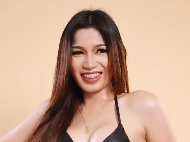 DaphneyLove with long brown hair, smiling with shiny teeth, wearing a black halter top, showing her tits, and a gold necklace against a plain beige background.