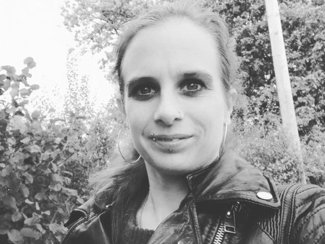 Black and white selfie of LadyChantale-hot with a nose ring, lip piercing, hoop earrings, and a leather jacket, standing outdoors with leafy bushes in the background.