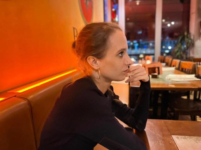 LadyChantale-hot with brown hair in a bun, wearing a black top, sitting in a restaurant, resting her chin on her hand, looking to the side, with gold earrings.