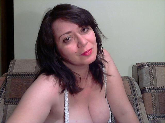 PerkyBoobsMature mit schulterlangem, schwarzem Haar, rosa Lippenstift und einem süßen Lächeln, trägt einen weißen Spitzen-BH, der ihre großen Brüste zeigt, und sitzt auf einem karierten Sofa..