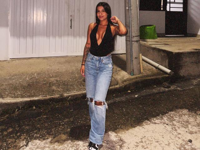 Thalia Ares tem cabelos longos e escuros, uma blusa preta com decote em V que mostra seus seios, e jeans azuis rasgados com um joelho desgastado. Ela está em pé em uma calçada suja à noite..