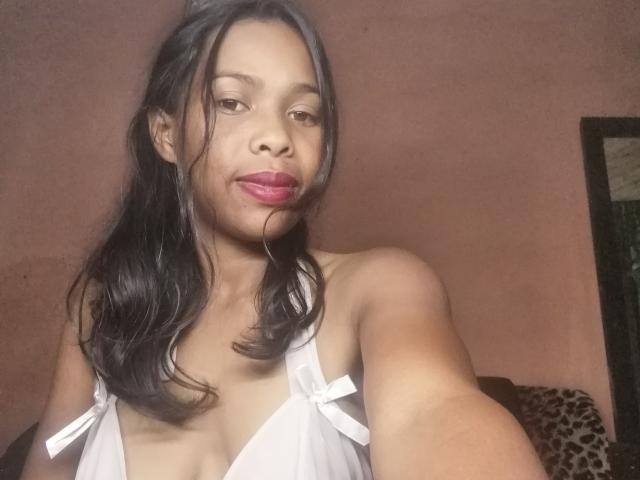 CathiaX with medium brown skin, black hair in a ponytail, pink lipstick, white halter top showing small tits, holding out arm for a selfie.