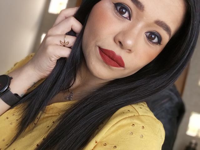 MistressVictoriaa has long black hair, red lipstick, and brown eyes. She's wearing a yellow shirt with small patterns, a black watch, and a ring on her right hand.