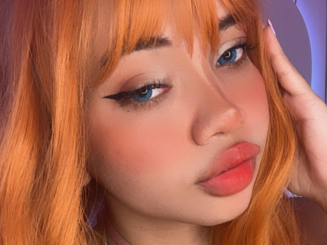 LiliMays has orange hair, blue eyes, and pink lips with black winged eyeliner, touching her head with her right hand. Her face is slightly tilted to the right.