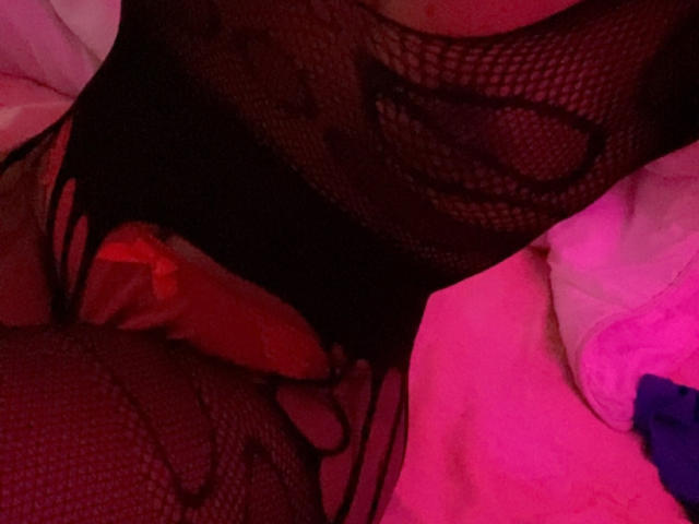 Het zwarte netlingerie van TamaraShow onthult haar roze verlichte vulva, met haar opgewekte, rood getinte penis die gedeeltelijk zichtbaar is door het gaas. Haar dijen zijn licht gespreid..