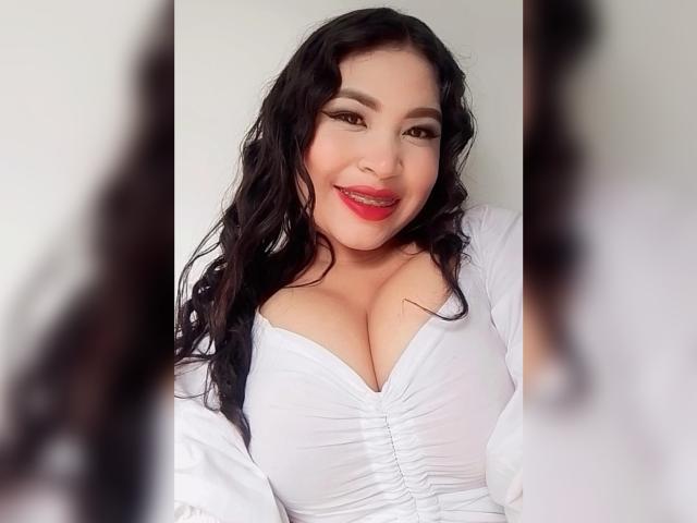 OliviGrace, avec ses cheveux noirs ondulés, son rouge à lèvres rouge et son haut blanc plissé, qui met en valeur de grandes, rondes et généreuses seins, sourit à l'appareil photo. Son reflet est flou des deux côtés..