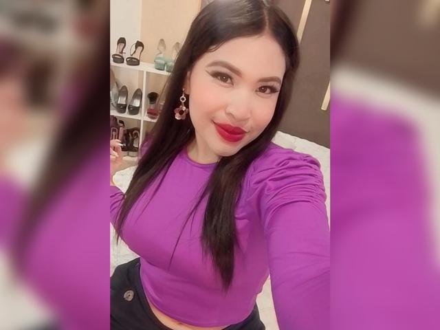 OliviGrace with long black hair, purple top, red lipstick, and earrings, showing off her tits in a selfie. Her reflection is on the right.