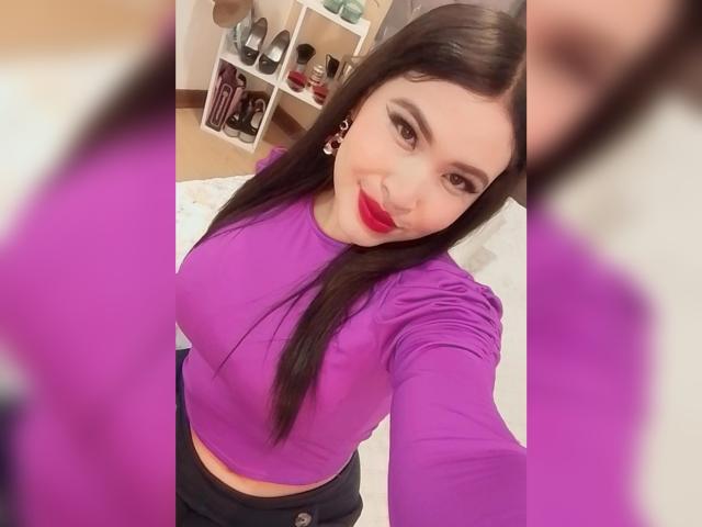 OliviGrace with long black hair, purple long-sleeve top, red lipstick, and dark pants, showing off her small, perky tits in a selfie. She's smiling and holding the camera.