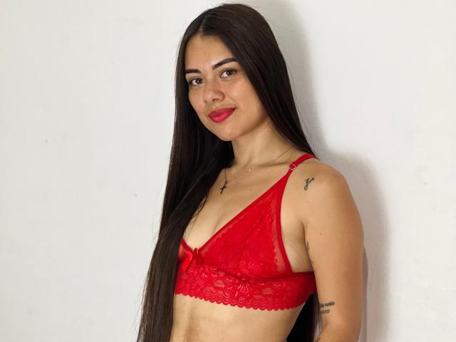 CelesteNoir with long dark hair, red lace bra, small tits, tattoo on left shoulder, tattoo on right arm, smiling, red lipstick, standing against white wall.