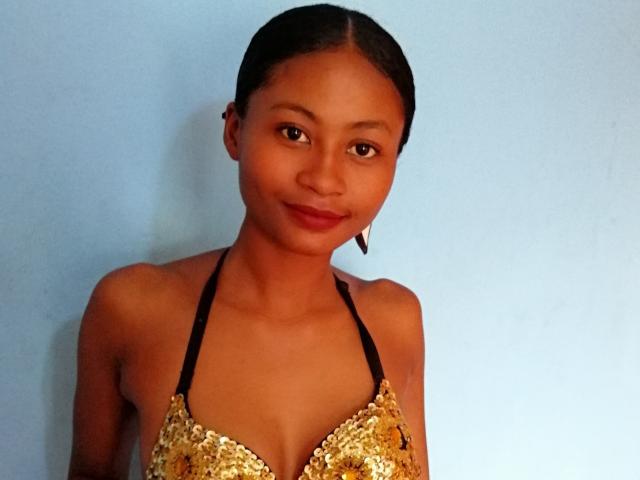 Cayrah with dark brown skin, small boobs, and a cute smile, wearing a gold sequined halter top, has black hair in a slick back, and small earrings.