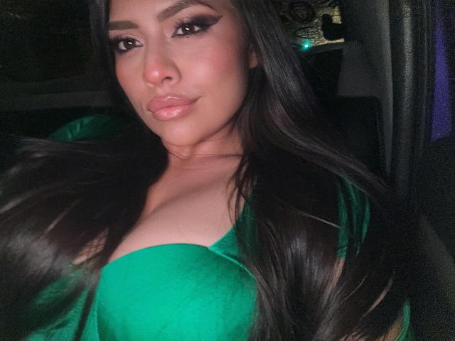 BellaSmithX69 with long, glossy black hair, green velvet top, showing her big, shiny tits, full lips, and dark, thick eyebrows, in a dimly lit car.