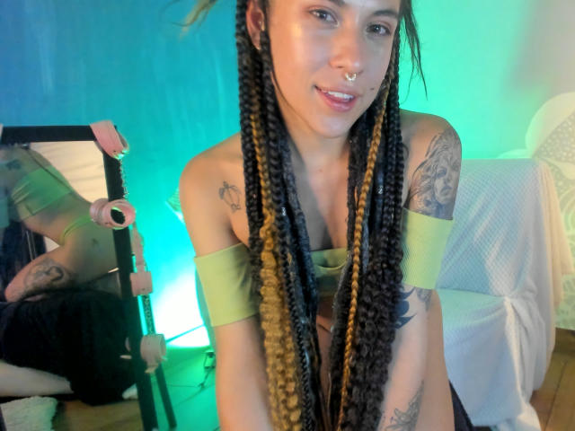 HellenK with long braided black and blonde hair, green off-shoulder top, tattoos, nose ring, and smiling, showing small tits and a cute face, in a softly lit room.