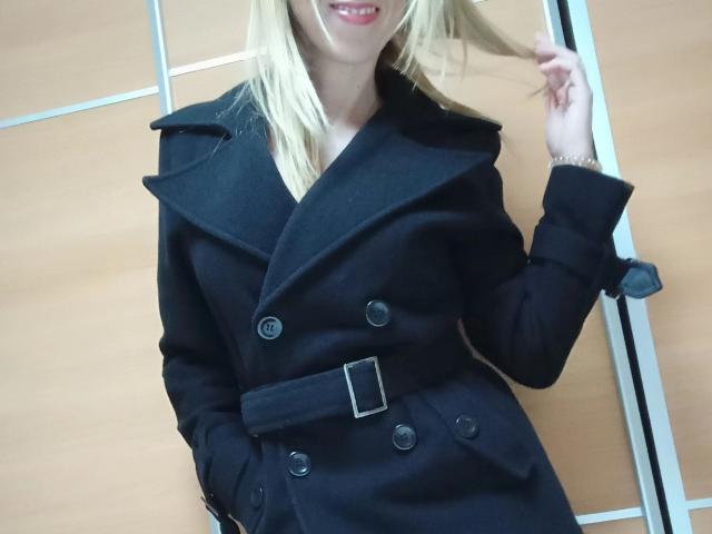 LaraCordoba-hot wearing a black, double-buttoned, belted coat with a wide collar, her blonde hair in hand, smiling with red lipstick, showing her chest.