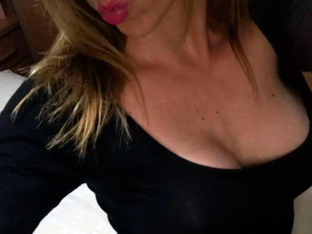 LaraCordoba-hot with pink lipstick, blonde hair, black top, showing her big boobs and cleavage. She's got a few moles on her chest. Her tits are prominent.