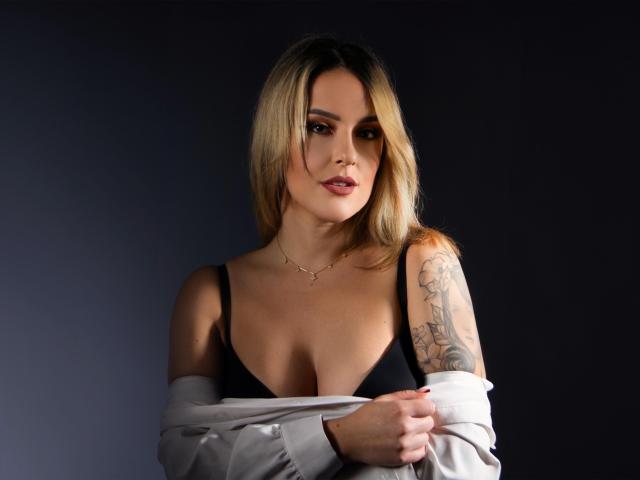 MollyPretty with blonde hair, black bra, and tattooed arm, holding off white shirt, exposing her tits, and wearing a gold necklace, against a dark background.