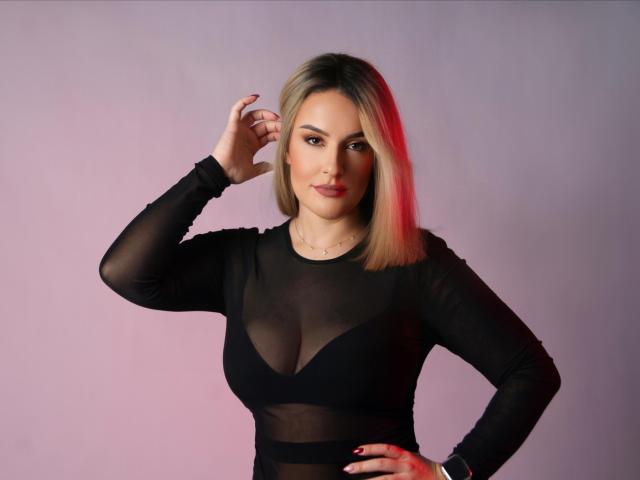 MollyPretty with blonde bob, wearing sheer black top, showing tits through mesh, hand on hip, looking at camera, red highlight in hair, confident expression.