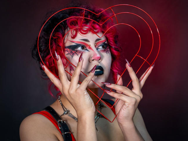 GothVioleth, avec ses cheveux rose-rouge bouclés, son rouge à lèvres noir, son fil métallique en forme de cœur rouge, son maquillage d'yeux en forme de toile d'araignée, ses ongles roses longs, ses bretelles noires et sa chaîne, tenant un cadre en forme de cœur, avec une expression séduisante..