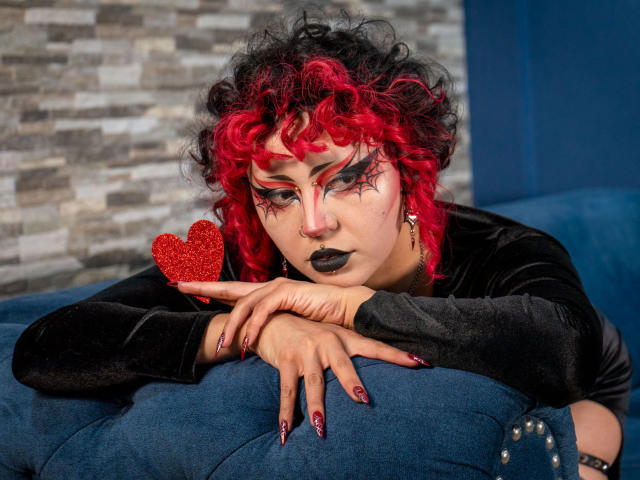 GothVioleth, avec des cheveux bouclés roux et noirs, du maquillage noir, tenant un cœur rouge, un haut en velours noir, du rouge à lèvres foncé, des ongles rouges, appuyée sur un canapé bleu, regardant vers le bas..