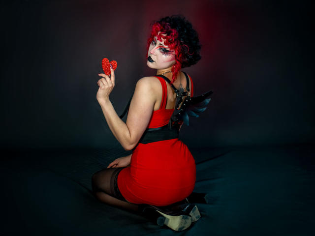 GothVioleth com cabelo ruivo e preto encaracolado, vestido vermelho, asas pretas, segurando um coração vermelho, batom preto, ajoelhada, mostrando o bumbum, usando meias pretas, saltos brancos, fundo escuro..