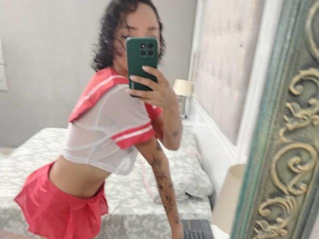 SilvanaXOwen vestida con un conjunto transparente rojo y blanco de colegiala, mostrando sus pequeños senos y falda roja, con un tatuaje en el brazo, tomando una selfie con un teléfono verde..