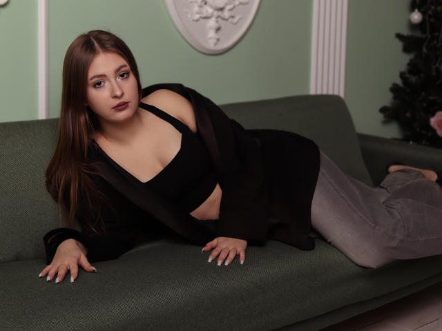 AdelPearson has brown hair, fair skin, and small tits. She's wearing a black top, grey pants, and a black jacket, lying on a green couch with a relaxed expression.