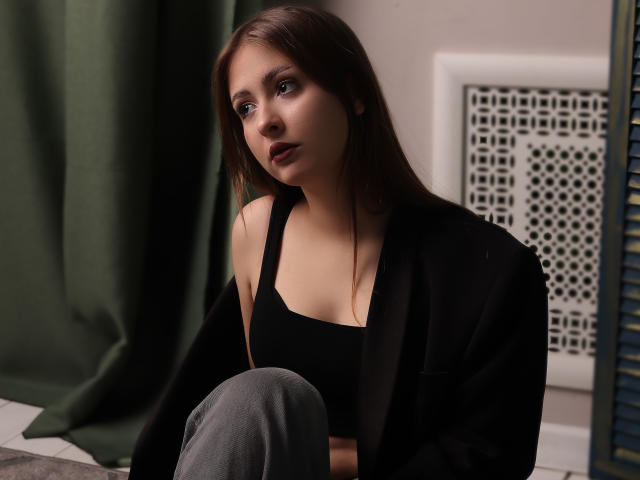 AdelPearson with long brown hair, wearing a black tank top and open black shirt, gray pants, looking slightly up with a subtle, serious expression, in a dimly lit room.