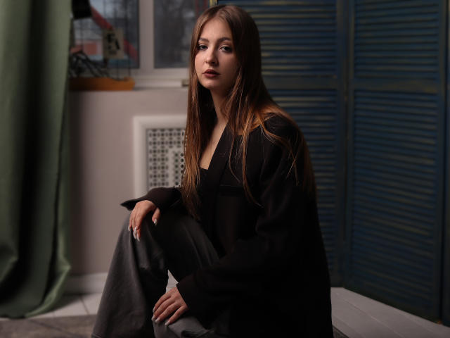 AdelPearson with long brown hair, wearing a black shirt and gray pants, sitting with a serious expression, blue shutters in the background, and green curtain on the left.