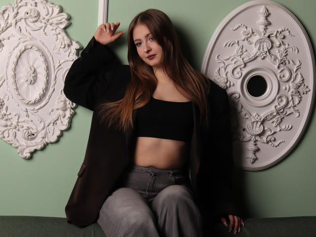 AdelPearson with long brown hair, black crop top showing her tits, grey pants, and black jacket, leaning against a green wall with ornate white decorations.