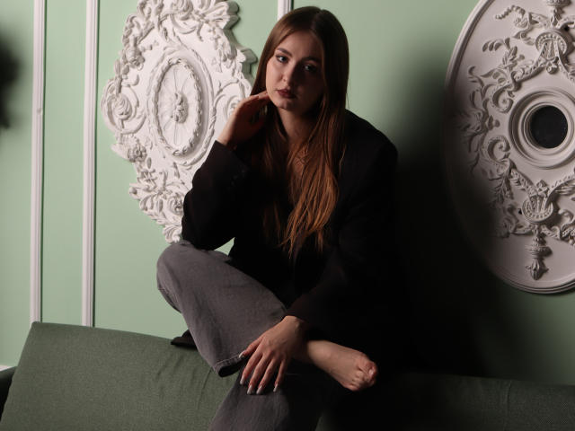 AdelPearson with long brown hair, sitting on green couch, wearing black blazer and gray pants, hand on cheek, looking at camera, bare feet, ornate white wall decor.